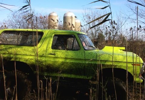 1978 Ford Bronco - Side profile of a lime green full-size Ford Bronco streaked with dried mud, sitting in tall reeds and brush with a lifted stance and knobby…