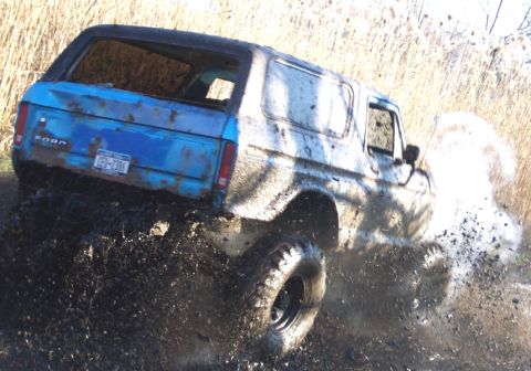 1978 Ford Bronco - Rear three-quarter view of a blue full-size Ford Bronco with a removable hardtop tearing through a mud pit, its passenger side plastered wh…