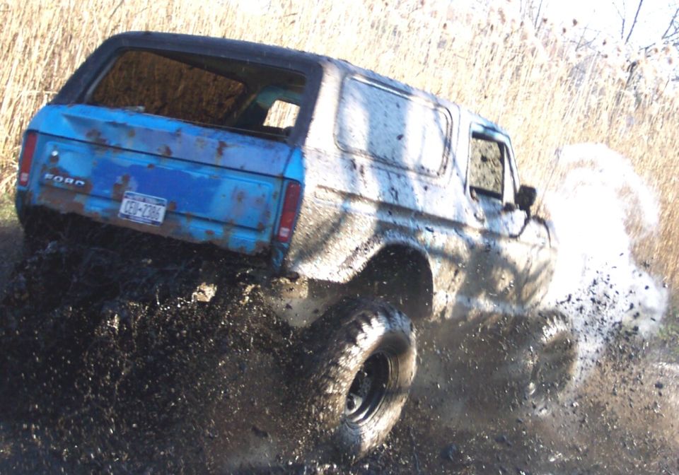 Blue full-size Ford Bronco caked in mud running a bog, rear three-quarter view