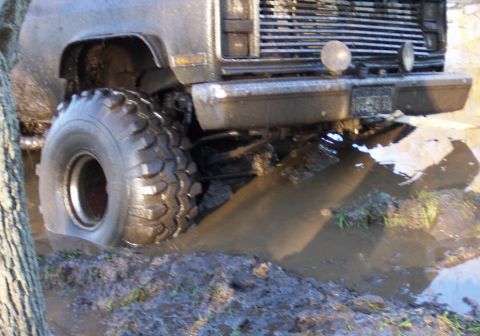 1978 Ford Bronco - Low close-up of the front corner of a square-body pickup sunk in a muddy water hole, showing the chrome bumper, stacked grille, round auxil…
