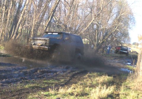 1978 Ford Bronco - A dark square-body Chevrolet Blazer-style two-door SUV blasts through a mud pit, slinging spray from all four tires as it runs a rutted tra…