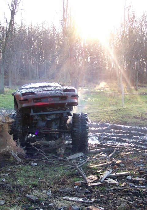 1978 Ford Bronco - Rear view of a red Chevrolet coupe body perched on a tall tube-frame 4x4 chassis, backlit by low sun as it crawls over logs and rubble at t…