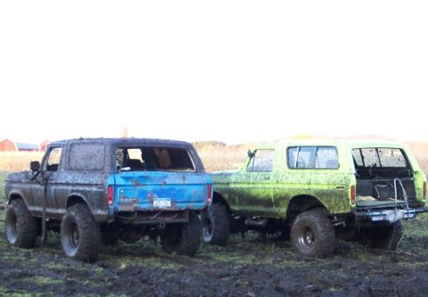 1978 Ford Bronco - Two lifted full-size Ford Broncos parked side by side in a muddy field, both splattered from a bog run: one wearing faded blue and gray pai…