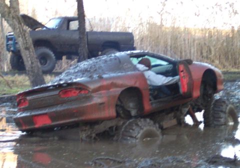 1978 Ford Bronco - A mud-covered red coupe fitted with oversized off-road tires and a lifted suspension sits stuck in a muddy pit, with a person visible insid…
