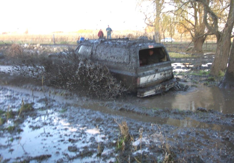 Mud-covered two-door SUV running a muddy bog with spectators watching
