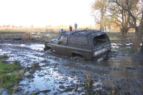 1978 Ford Bronco - A square-body Chevrolet Blazer/K5-style SUV, completely plastered in gray mud, churns through a deep flooded bog pit with water and muck sp…
