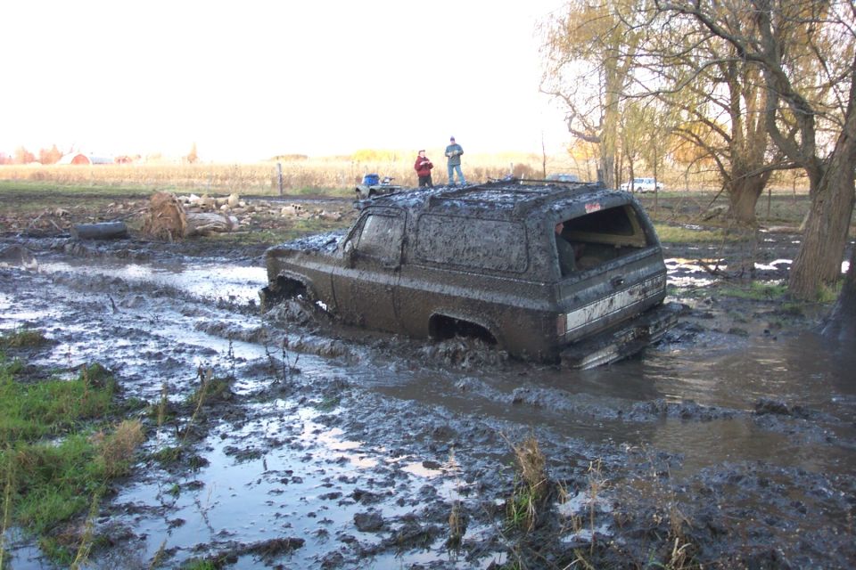Mud-plastered square-body SUV churning through a deep flooded bog pit