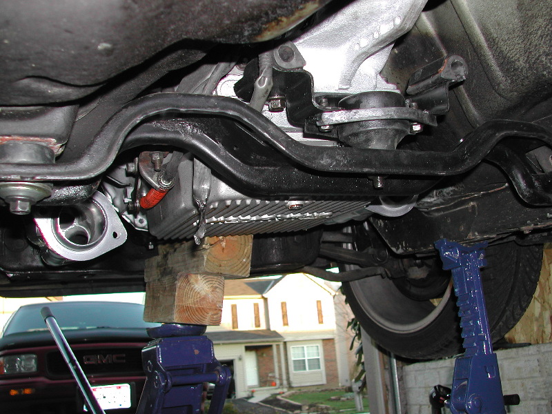 Underside view of a vehicle on jack stands, showing suspension and exhaust components. A GMC truck is visible in the background