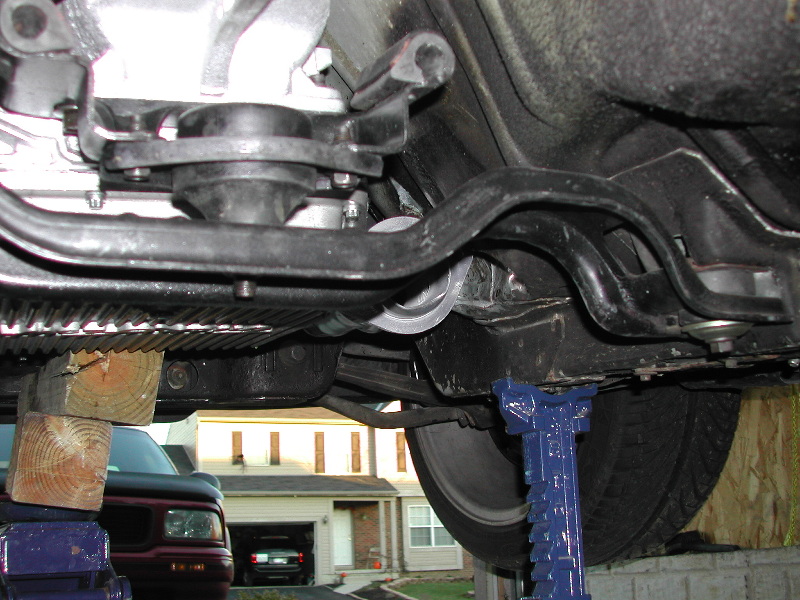 Undercarriage of a vehicle supported by jack stands and wooden blocks, with a red pickup truck and a house visible in the background