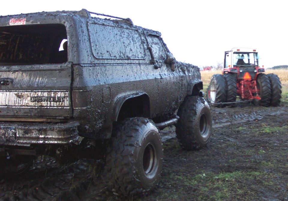 Mud-covered Chevrolet K5 Blazer, rear view, with a tractor in the background