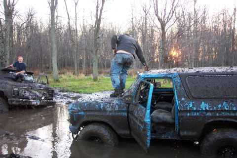 1978 Ford Bronco - A blue full-size Ford Bronco, doors off and cab caked in gray mud, sits axle-deep in a water-filled bog pit while a man in jeans stands on…