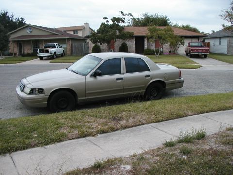 1999 Ford Crown Victoria - Gold Ford Crown Victoria parked on street with two pickup trucks in background driveways.