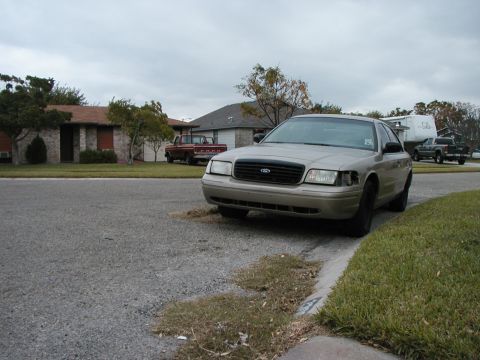 1999 Ford Crown Victoria - A tan Ford Crown Victoria is parked on the side of the road, with houses and other vehicles visible in the background.