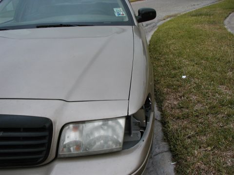 1999 Ford Crown Victoria - A tan sedan is parked on the side of the road, showing damage to its front passenger side headlight.
