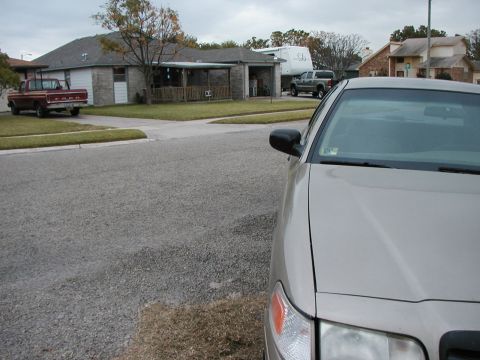 1999 Ford Crown Victoria - A tan sedan is parked in the foreground, with a red pickup truck and a gray pickup truck visible in the background of a residential street.