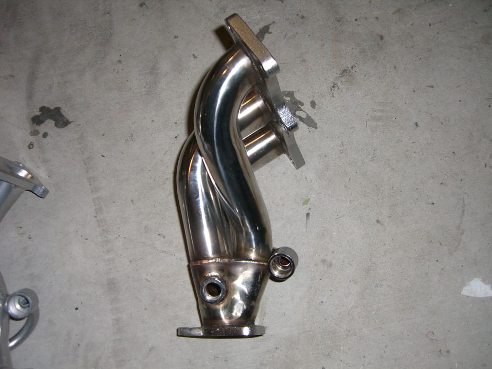 Shiny metal exhaust manifold with four pipes merging into one, resting on a concrete floor
