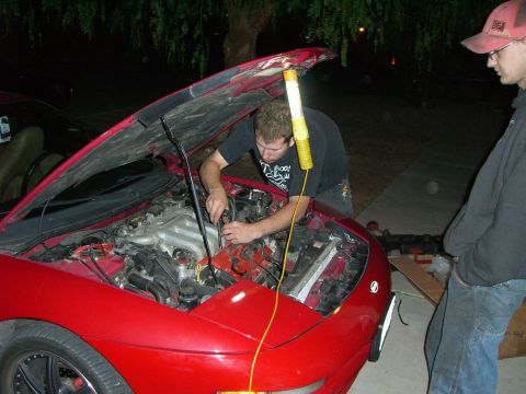1996 Ford Probe - Two men work on the engine of a red car at night, with the hood fully open.