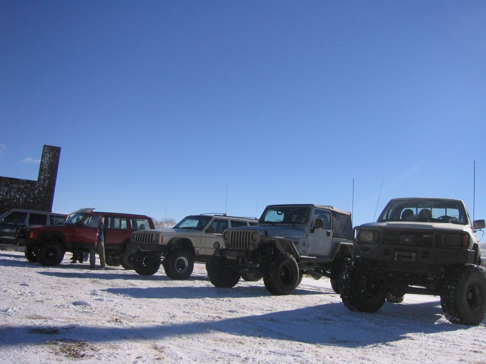 Several modified Jeeps and a pickup truck parked in a snowy field under a clear blue sky