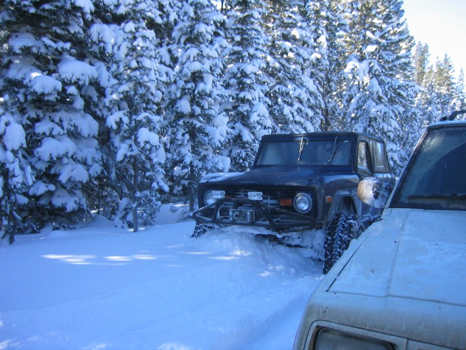 Dark blue Ford Bronco with winch in snow, front 3/4 view. Another vehicle is partially visible on the right