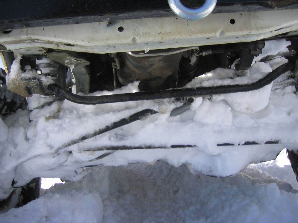 Undercarriage of a vehicle covered in snow, showing suspension components and a tow hook