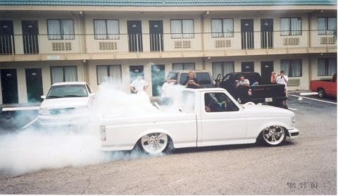 1986 Ford Bronco - A lowered white 1990s Ford F-150 regular-cab short-bed on polished billet wheels does a smoky burnout in a motel parking lot, tire smoke po…