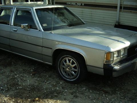 1991 Ford LTD Crown Victoria - A silver sedan with chrome wheels is parked outdoors next to a trailer.