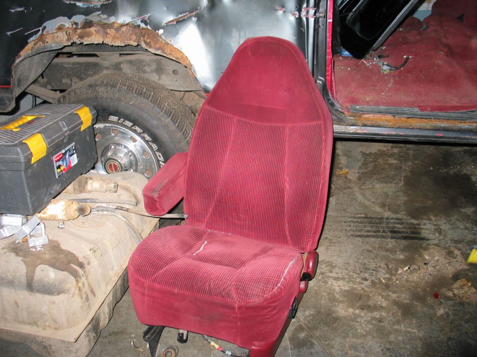 Red velour bucket seat in a vehicle with a rusty wheel well, fuel tank, and toolbox nearby