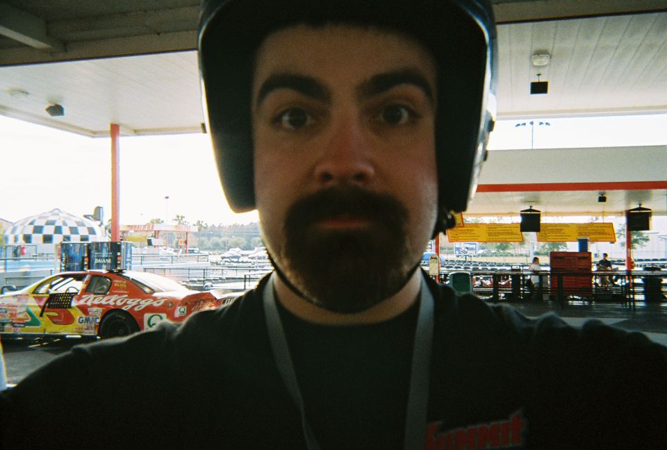 Bearded man in a black karting helmet at a go-kart track with a Kellogg's stock car behind him