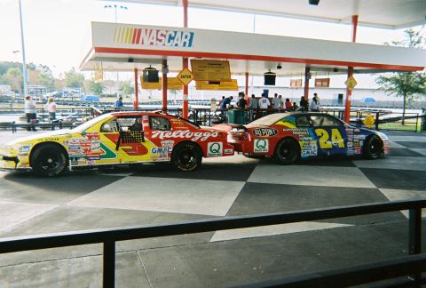 1991 Ford Probe - Two NASCAR race cars are parked under a canopy at what appears to be a race track or amusement park.