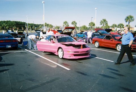 1991 Ford Probe - A pink sports car is displayed with its hood open at an outdoor car event, with several other vehicles present.