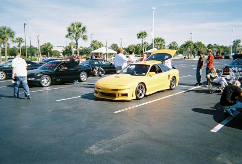 1991 Ford Probe - A yellow sports car with a custom body kit is parked in a lot with its trunk open, surrounded by other vehicles and people.