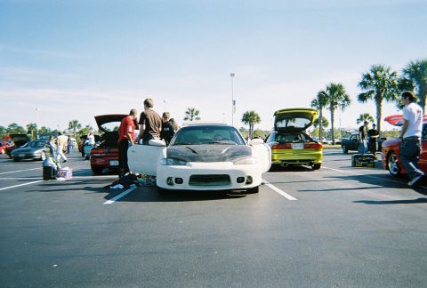 1991 Ford Probe - Several cars are gathered in a parking lot on a sunny day, with people milling around them.