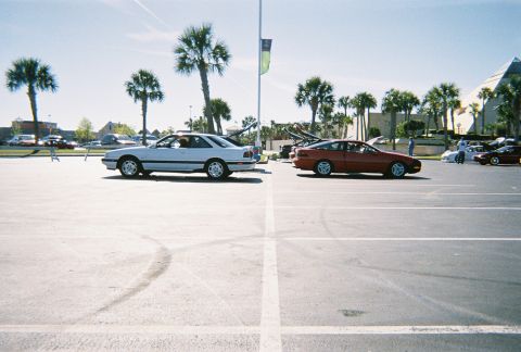 1991 Ford Probe - Two coupes are parked side-by-side in a parking lot under a bright, sunny sky with palm trees.