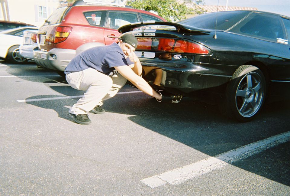 Man crouches to inspect exhaust pipe of a black 1991 Ford Probe, red SUV parked behind