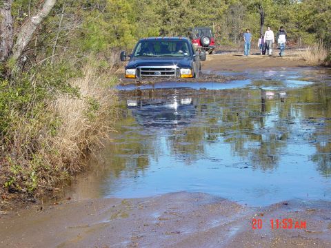 1995 Dodge 1500 - A dark-colored Ford F-Series truck navigates a deep water crossing on a dirt road, with a Jeep and onlookers in the background.