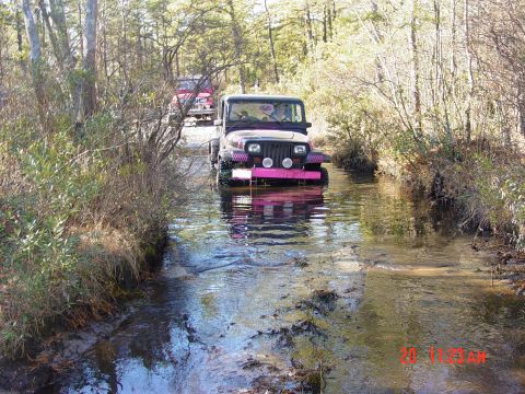 1995 Dodge 1500 - Two off-road vehicles navigate a shallow stream surrounded by trees.
