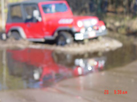 1995 Dodge 1500 - A red Jeep Wrangler navigates through a muddy water crossing, its reflection visible in the water.