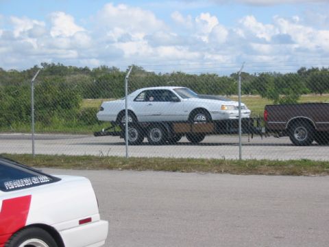 1997 Ford Crown Victoria - A white coupe from the 1990s is transported on a trailer, with a brown pickup truck visible behind it.