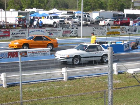 1997 Ford Crown Victoria - Two Ford Mustangs, one orange and one white, compete on a drag strip.