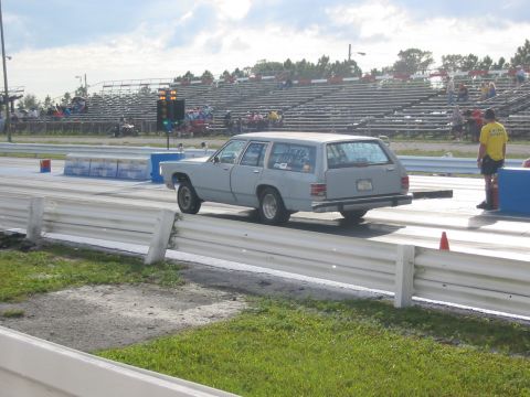 1997 Ford Crown Victoria - A station wagon is on the drag strip with spectators in the stands and a track official nearby.