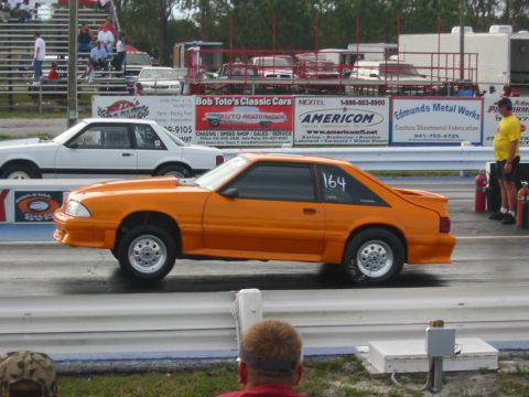 1997 Ford Crown Victoria - A drag race is underway with an orange Ford Mustang in the foreground and a white Ford sedan in the background.