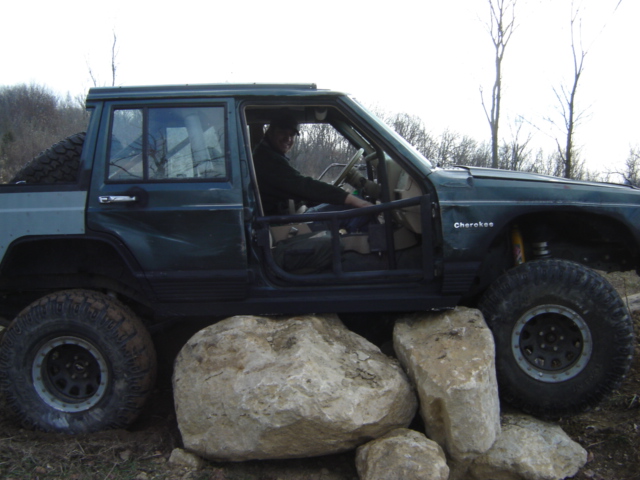 Green Jeep Cherokee with off-road modifications, driver visible, positioned over large rocks