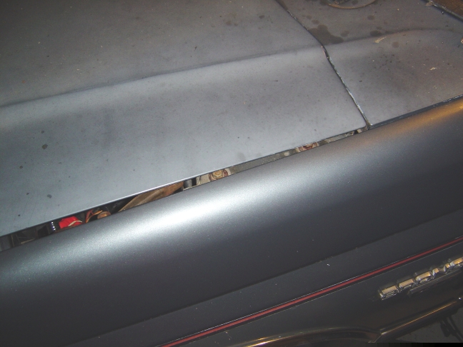 close-up of a car's hood seam, showing a gap with some engine parts visible underneath. A chrome trim piece runs along the fender