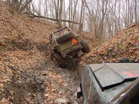 1992 Ford Bronco - Off-road vehicles struggle through a muddy, wooded trail with one vehicle tilted precariously.