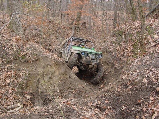 Green off-road vehicle with roll cage descending a muddy, wooded ravine