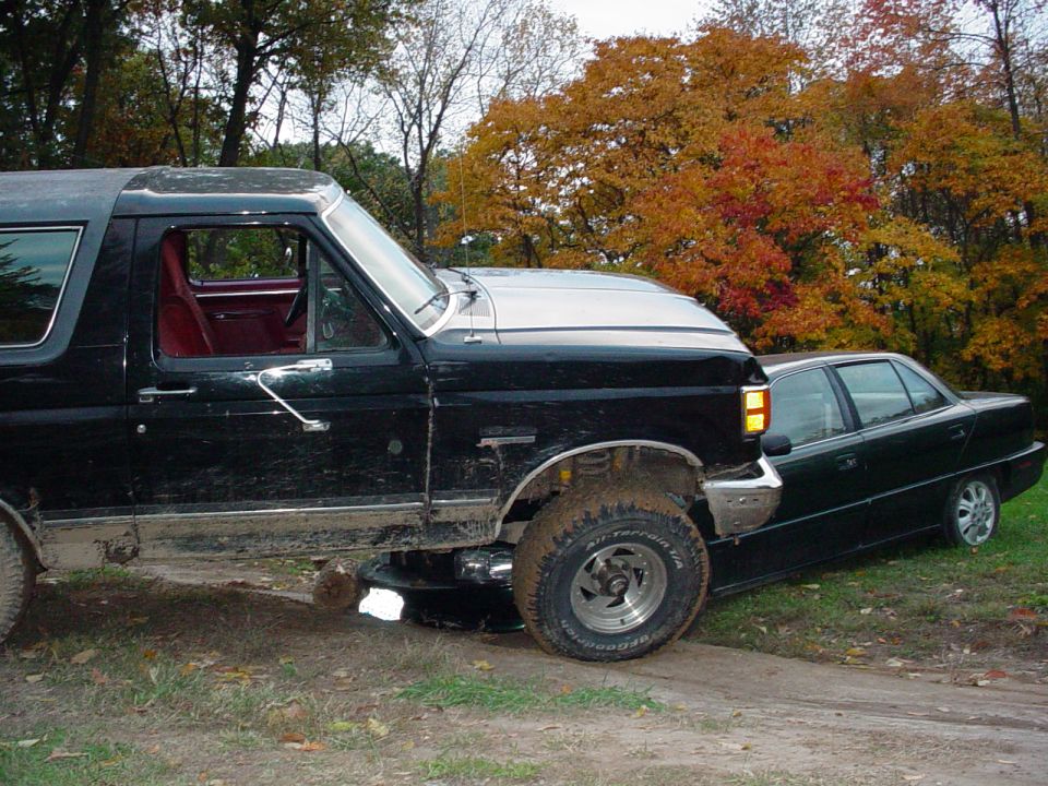 1988 Ford Bronco parked next to a dark sedan on a dirt path with fall foliage in the background