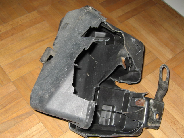 Damaged black plastic car part with metal bracket attached, lying on a wood floor