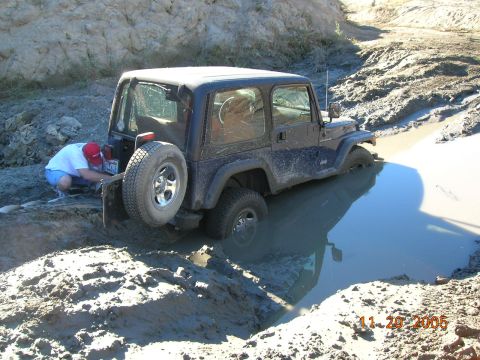 1985 Ford Bronco - Jeep Wrangler is stuck in a muddy water hole, a person is attempting to fix the rear bumper.
