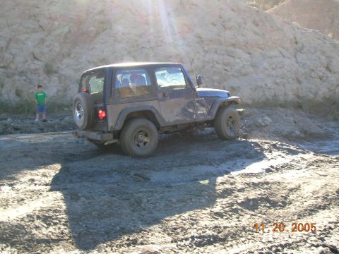 1985 Ford Bronco - A Jeep Wrangler navigates a muddy trail with a rocky hillside in the background.