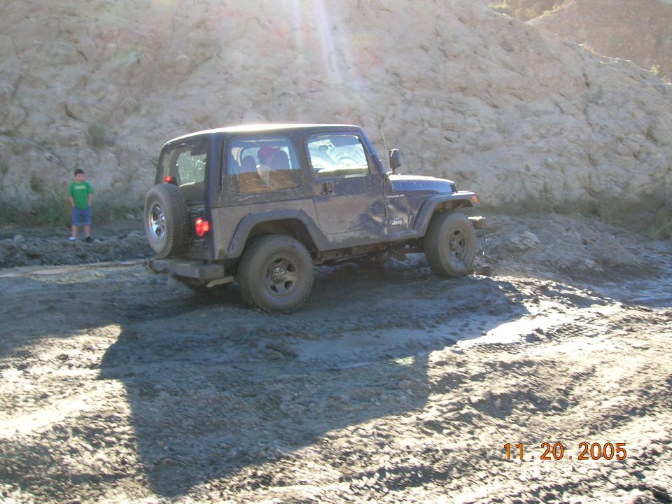 Dark blue Jeep Wrangler on muddy terrain, spare tire on rear door, sun glare